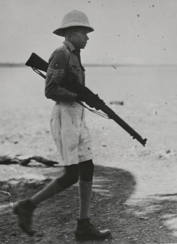 Louis Munkara walking on a beach holding a rifle.