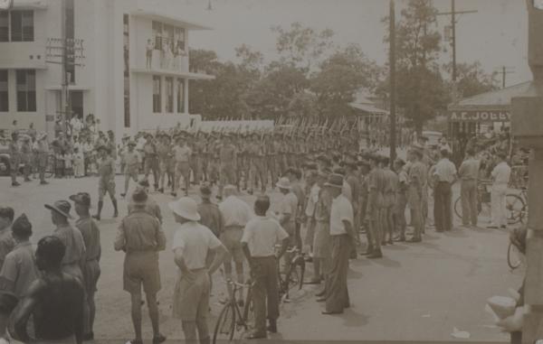 Soldiers marching down a street with a crowd gathered watching.