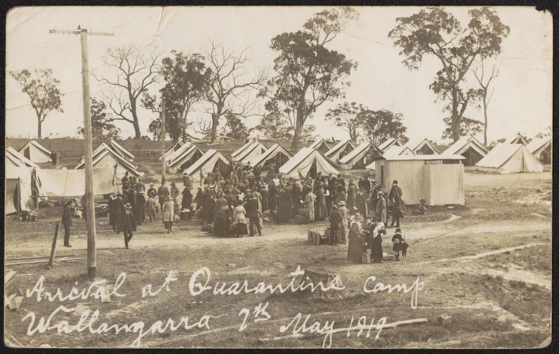 Large group of men and women arrive at camp of tents, with toilet block at right, power pole at left and gum trees in the distance.