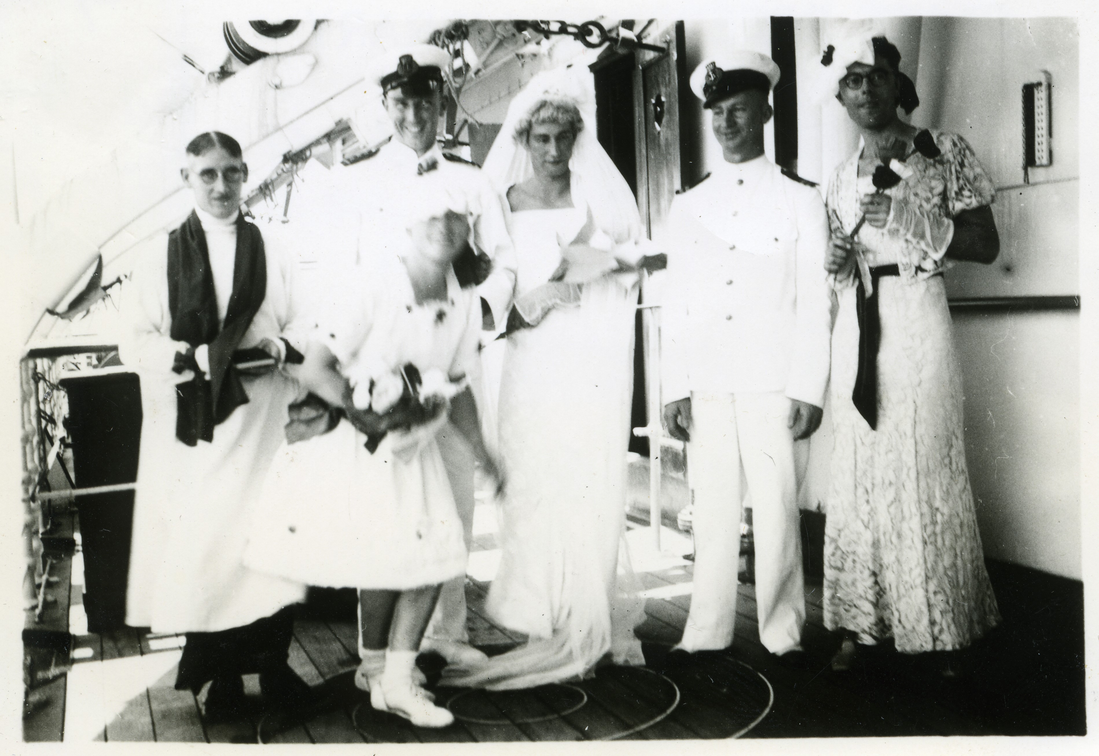 Six men on the deck of a ship dressed as a wedding party including pastor, bridesmaids and bride. 