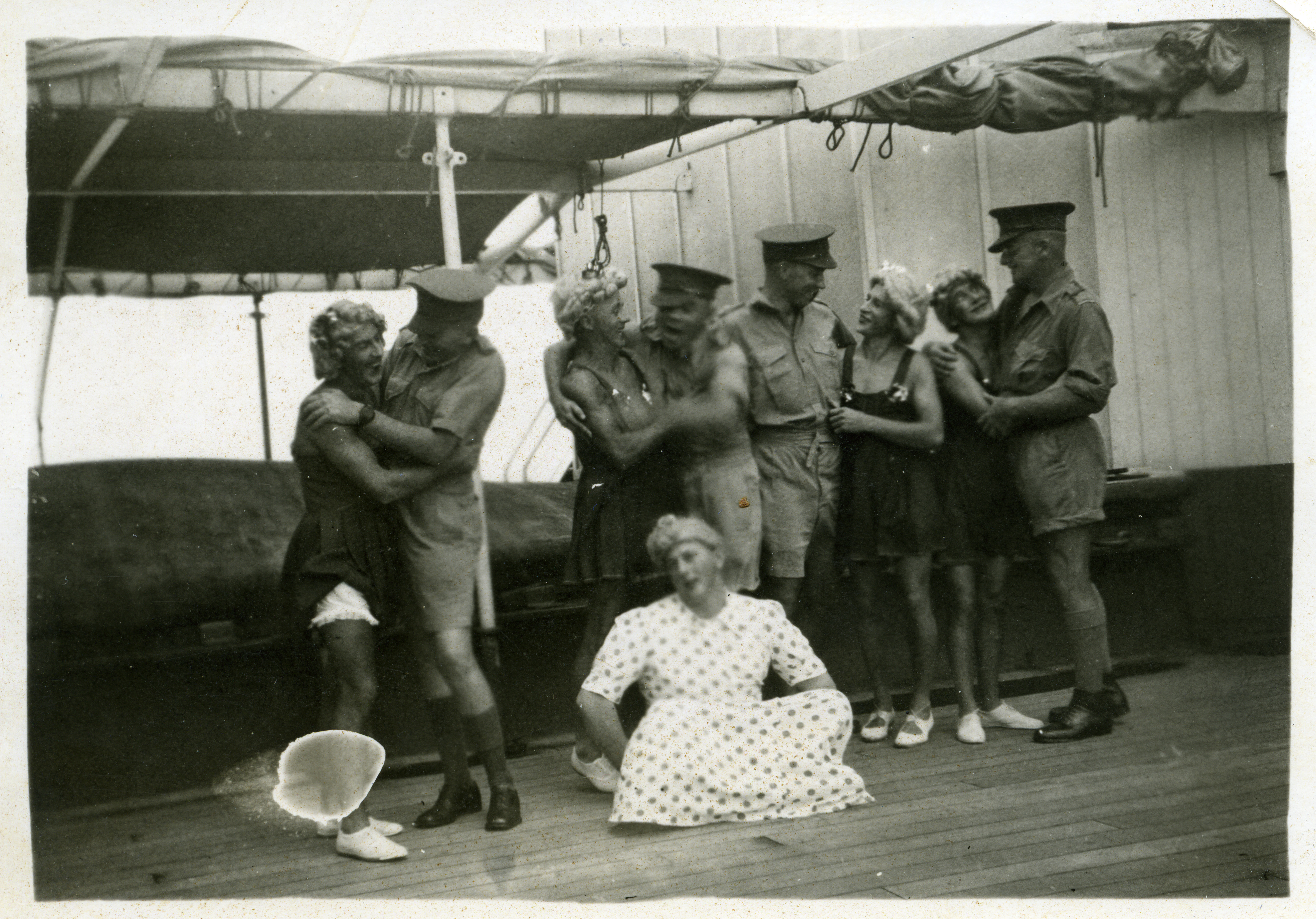 Four “couples” comprised of officers in uniform with their cross-dressing “girls”. A man in a white spotted dress sits at their feet.