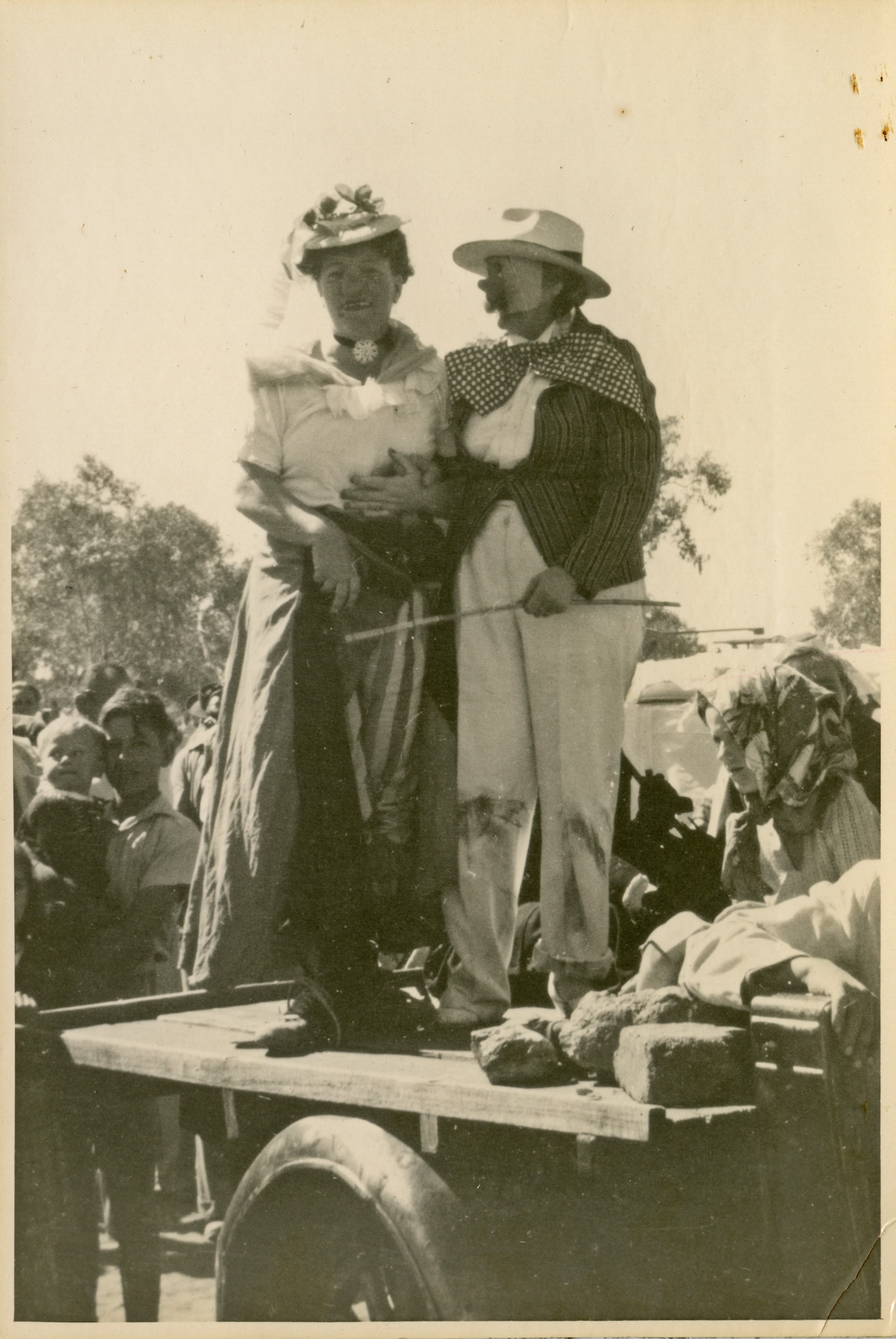 A cross-dressing, masked couple standing on the back of a truck in front of a crowd. 